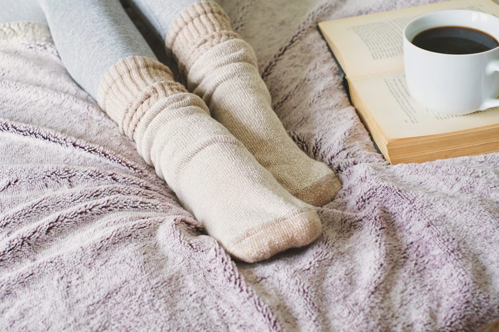 Young woman suffering from Raynaud's disease wearing grey tights and cozy socks sitting on her bed with a book and coffee