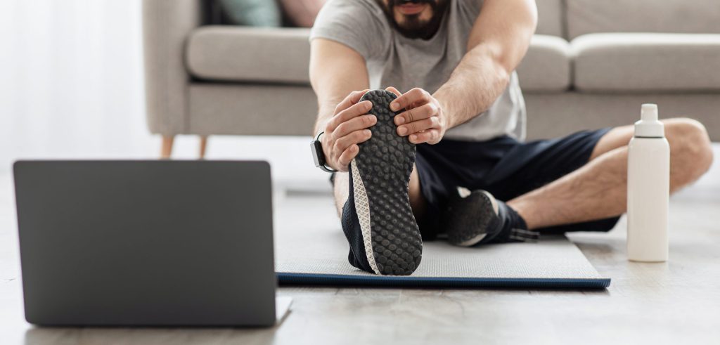 Arabian bearded man Exercising at Home and doing stretching in living room interior, on floor, looking at laptop with bottle of water on floor.