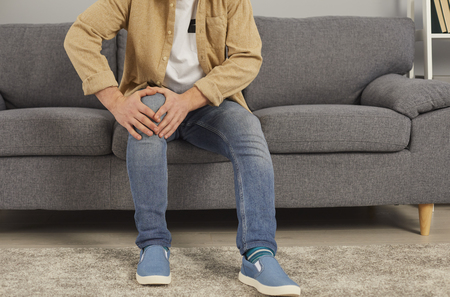 Man who has arthritis or rheumatism sitting on sofa and touching his painful knee