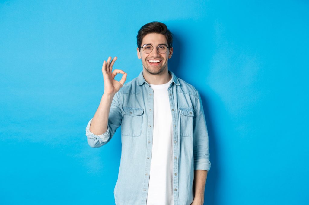 Attractive young man wearing glasses and casual clothes, showing ok good sign in approval, like something, standing against blue background.