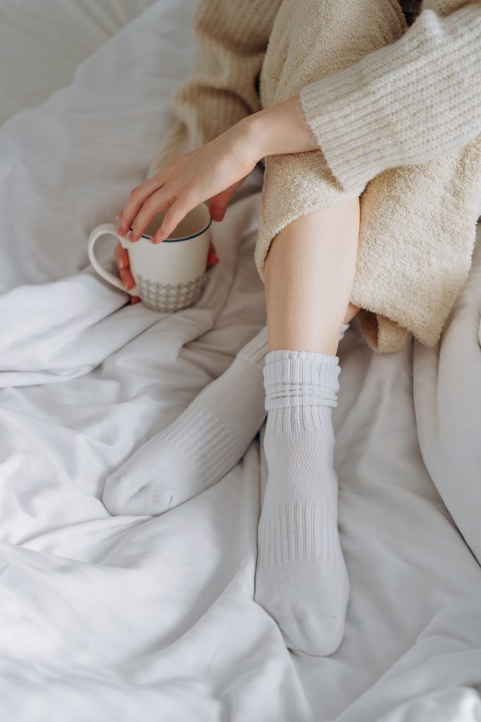Woman in White Sweater Holding White Ceramic Mug. Cold feet in socks.