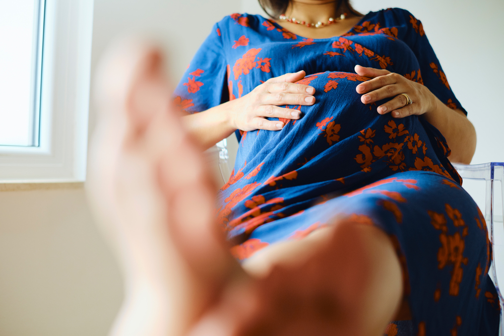 Barefoot young pregnant woman sitting on chair with feet up and hands on stomach. Feet swelling during pregnancy concept.