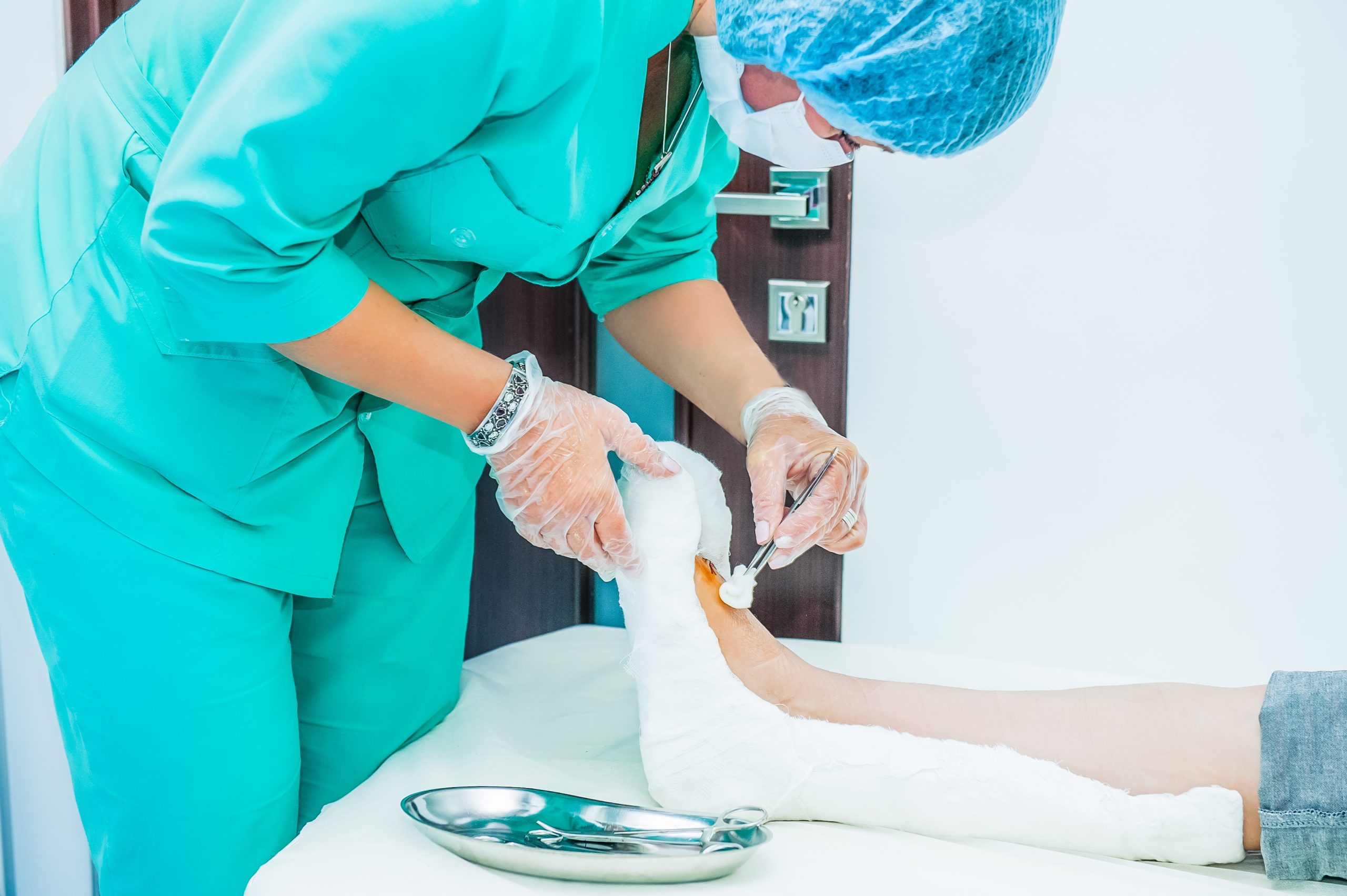 A podiatrist (feet doctor) wearing a green scrub and treating a foot ulcer on the patient's leg in a clinic room