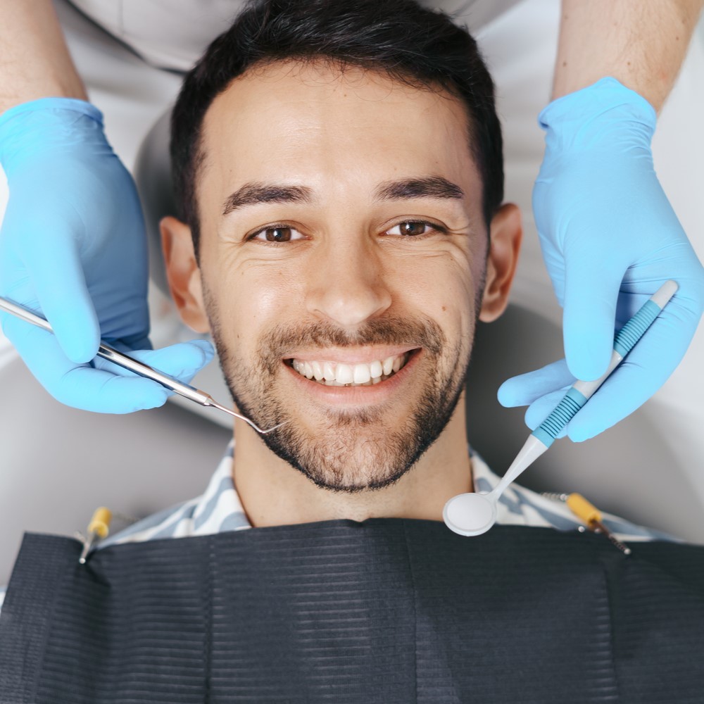 Smiling young man sitting in dentist chair while doctor examining his teeth. Regular dentist visits concept.