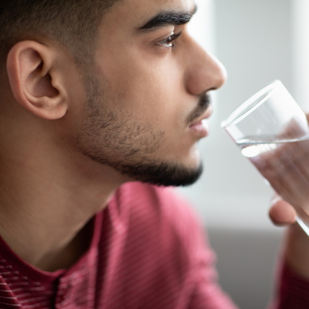 Hydration, healthy lifestyle, water drinking concept. Closeup of young bearded Arab man holding glass, drinking fresh water at home, side view, panorama, looking at copy space. Dry mouth during fasting concept.